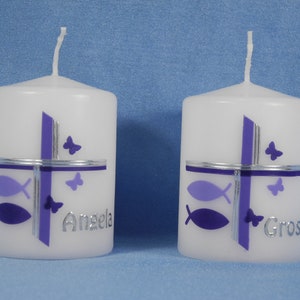 K&ouml;nnte beinhalten: Zwei wei&szlig;e Kerzen mit violetten und silbernen Designs. Die Kerzen haben ein Kreuz, Fische und Schmetterlinge. Eine Kerze sagt "Angela" und die andere sagt "Grosi".