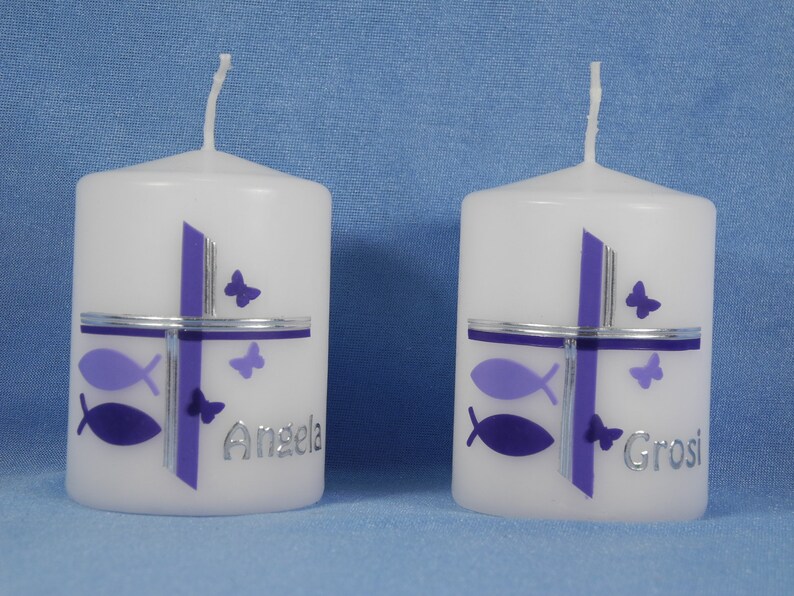 K&ouml;nnte beinhalten: Zwei wei&szlig;e Kerzen mit violetten und silbernen Designs. Die Kerzen haben ein Kreuz, Fische und Schmetterlinge. Eine Kerze sagt "Angela" und die andere sagt "Grosi".