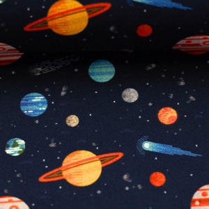 Könnte beinhalten: Dunkelblauer Stoff mit einem sich wiederholenden Muster aus Planeten, Sternen und Kometen. Die Planeten sind orange, blau und rot mit Ringen und Streifen. Weiße Sterne und blaue Kometen sind über dem Stoff verstreut.