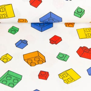 Könnte beinhalten: Weißer Stoff mit einem sich wiederholenden Muster aus bunten Spielzeugbausteinen. Die Bausteine sind in verschiedenen Farben gehalten, darunter Rot, Gelb, Blau, Grün und Orange. Die Bausteine sind über den Stoff verstreut.
