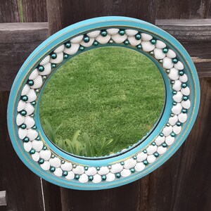 May include: A teal blue round mirror with a decorative frame made of white seashells and teal blue beads. The mirror reflects a green grassy lawn.