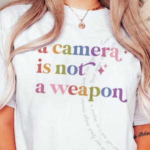 以下が含まれることがあります： 前面に「a camera is not a weapon」というマルチカラーのテキストがプリントされた、白い半袖Tシャツ。文字には赤、黄、緑、青、紫などのさまざまな色が使われており、小さな星のシンボルがアクセントとして添えられています。