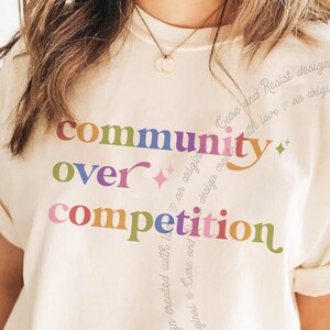 May include: A cream-colored short-sleeve t-shirt featuring the multicolored text "community over competition" printed on the front. The design includes small star icons and a swirling script pattern that reads "Care and Resist design created with love an original".