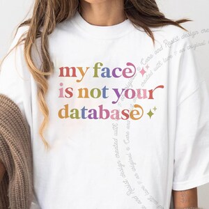 May include: A white short-sleeved t-shirt featuring the multicolored text "my face is not your database" printed on the chest. The design includes small star icons and stylized lettering in shades of red, blue, orange, and purple.