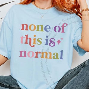 May include: A light blue short-sleeve t-shirt featuring the multicoloured text "none of this is normal" printed on the front in a retro-style font. The design includes a small four-pointed star icon next to the word "is" in a vibrant colour palette.