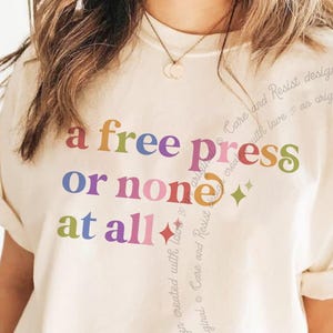 Puede incluir: Una camiseta de manga corta de color crema con el texto multicolor 'a free press or none at all' impreso en la parte delantera. El diseño incluye pequeños iconos de estrellas y un texto tenue y repetitivo que dice 'Care and Resist design created with love an original'.