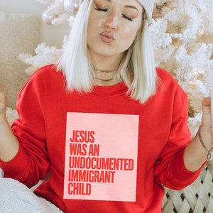 May include: A woman wearing a red sweatshirt with the text "JESUS WAS AN UNDOCUMENTED IMMIGRANT CHILD" in pink. She is holding a white mug with black speckles.