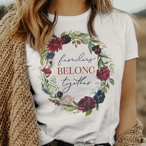 May include: White t-shirt with a floral wreath design and the text "families belong together".