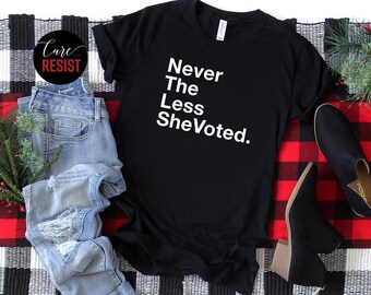 May include: Black t-shirt with white text that reads "Never The Less She Voted."