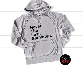 May include: A gray hooded sweatshirt with the text "Never The Less She Voted." printed on the front. The sweatshirt has a distressed look and is made of a soft, comfortable fabric.