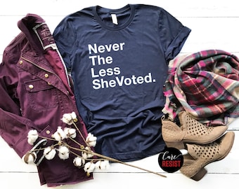 May include: Navy blue t-shirt with the text "Never The Less She Voted." A burgundy jacket, a plaid scarf, and tan ankle boots are arranged with the shirt. A sprig of cotton adds a natural element to the display. The shirt is a casual wear item.