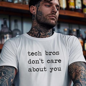 Puede incluir: Una camiseta blanca con el texto "tech bros don't care about you" impreso en la parte delantera. La camiseta la lleva puesta una persona con tatuajes en los brazos.