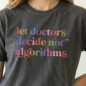 May include: A charcoal gray short-sleeve t-shirt featuring the multi-colored text "let doctors decide not algorithms" printed on the front. The shirt has a crew neckline and a relaxed fit, paired with blue denim jeans visible at the waistline.