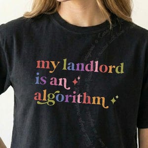 May include: A black short-sleeved t-shirt featuring the multicolored text "my landlord is an algorithm" printed on the front. The design includes small star-shaped accents and a subtle, curved watermark of text running vertically across the shirt.