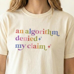 May include: A cream-colored short-sleeve t-shirt featuring the text "an algorithm denied my claim" printed in a multi-colored, wavy font. The design includes small four-pointed star icons interspersed between the words in various pastel hues.