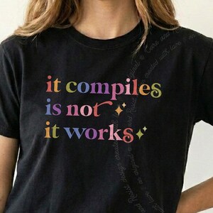 May include: A black short-sleeved t-shirt featuring the multi-colored text "it compiles is not it works" printed on the front. The design includes small star-shaped accents and subtle, faint cursive text swirling in the background behind the main graphic.