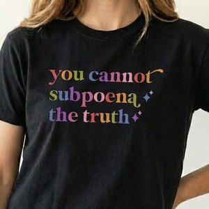 Può includere: Una t-shirt nera a maniche corte con il testo "you cannot subpoena the truth" stampato in un carattere sfumato multicolore. Il design include piccole icone a forma di stella a quattro punte intervallate tra le parole sul davanti.