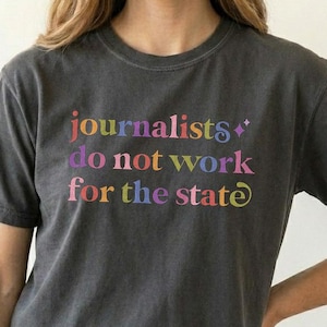 Può includere: Una t-shirt a maniche corte grigio antracite con il testo multicolore "journalists do not work for the state" stampato sul davanti. Le lettere appaiono in tonalità rosa, arancione, giallo, blu e viola con una piccola icona a stella.