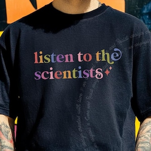 May include: A black short-sleeved t-shirt featuring the text "listen to the scientists" printed in a multi-colored, stylized font across the chest. The design includes small star-shaped accents and subtle, faint text flowing vertically down the shirt.