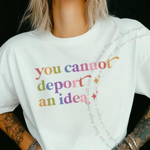 May include: A white short-sleeve t-shirt featuring the multicoloured text "you cannot deport an idea" printed on the front. The design includes a curved line of smaller, faint text and small star icons extending from the main phrase.