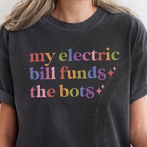 May include: A charcoal gray short-sleeve t-shirt featuring the multicolored text "my electric bill funds the bots" printed on the front. The design includes small four-pointed star icons in a retro-inspired font style with a crew neckline.