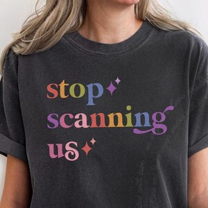 May include: A charcoal gray short-sleeve t-shirt featuring the multi-colored text "stop scanning us" printed on the front. The lettering is arranged in three lines with small four-pointed star icons in pink and purple accents near the text.
