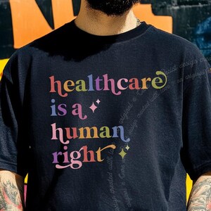 May include: A black short-sleeved t-shirt featuring the multicolored text "healthcare is a human right" printed on the front. The design includes small star-shaped accents and uses a stylized, flowing font with a gradient of colors.