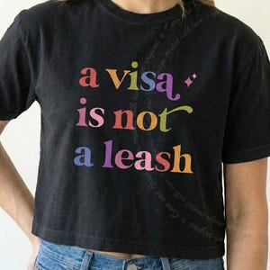 May include: A black short-sleeved t-shirt featuring the multicoloured text "a visa is not a leash" printed on the front. The shirt has a crew neckline and a straight hem, paired with blue denim jeans. The design uses various colours for the letters.