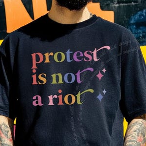 Puede incluir: Una camiseta negra de manga corta con el texto multicolor "protest is not a riot" impreso en la parte delantera. El diseño incluye pequeños iconos de estrellas de cuatro puntas en rosa y azul, con letras en tonos rosa, naranja y verde.