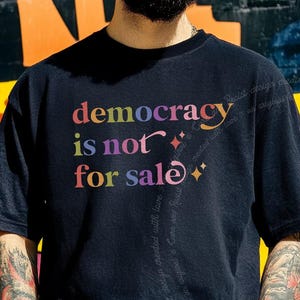 May include: A black short-sleeved t-shirt featuring the multicoloured text "democracy is not for sale" printed on the chest. The design includes small star-shaped accents and a swirling, faint watermark text pattern extending down the right side.
