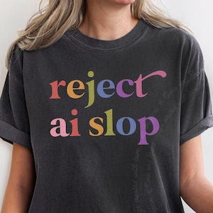 May include: A charcoal gray short-sleeve t-shirt featuring the text "reject ai slop" printed in a multi-colored, serif font. The letters are colored in shades of pink, yellow, green, blue, and purple, creating a vibrant contrast against the dark fabric.