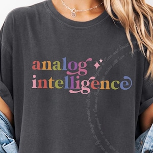 Może przedstawiać: Ciemnoszary t-shirt z krótkim rękawem z napisem "analog intelligence" wykonanym wielokolorową czcionką gradientową na klatce piersiowej. Koszulka jest założona pod jasnoniebieską kurtkę dżinsową z długimi rękawami i zapinanymi mankietami.