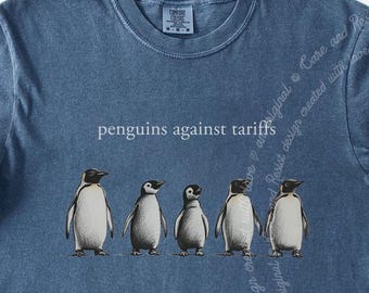 May include: Blue t-shirt with the phrase "penguins against tariffs" and a graphic of five penguins. The penguins are in black and white, standing in a row. The shirt has rolled-up sleeves.