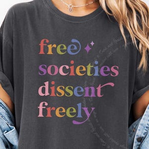Puede incluir: Una camiseta de manga corta gris carbón con el texto multicolor "free societies dissent freely" impreso en la parte delantera con una fuente estilizada, acompañada de un pequeño gráfico de estrella de cuatro puntas y una chaqueta vaquera azul claro.