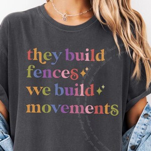 May include: A charcoal grey short-sleeve t-shirt featuring the multicoloured text "they build fences we build movements" with small star icons. The shirt is worn with a light blue denim jacket layered over the shoulders, showing a casual style.