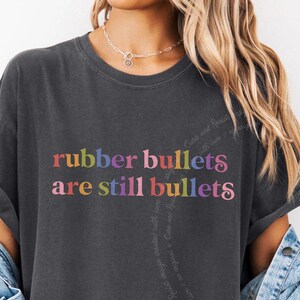 May include: A charcoal gray short-sleeve t-shirt featuring the text "rubber bullets are still bullets" printed in a multi-colored, rounded font across the chest. The shirt has a crew neckline and is worn with a light blue denim jacket.