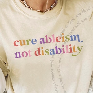 May include: A cream-colored crewneck t-shirt featuring the multicolored text "cure ableism not disability" printed across the chest. The design includes a subtle, repeating vertical script pattern that reads "Care and Resist design created with love an original" in a light gray tone.