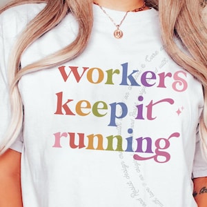 May include: A white short-sleeve t-shirt featuring the text "workers keep it running" printed in a multi-colored gradient font. The letters transition through shades of red, orange, yellow, green, blue, and purple across the three lines of text.