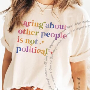 Op de afbeelding: Een wit T-shirt met de tekst "caring about other people is not political" gedrukt in regenboogkleuren.