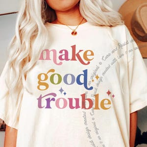 以下が含まれることがあります： カラフルな文字で「make good trouble」と書かれた白いTシャツ。文字はレトロなフォントで、縦に積み重ねられています。