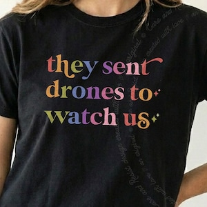 Puede incluir: Una camiseta negra de manga corta con el texto "they sent drones to watch us" impreso en una fuente de degradado multicolor. Las letras transicionan a través de tonos amarillos, naranjas, rosas, morados y azules, con dos pequeños iconos de estrellas de cuatro puntas.