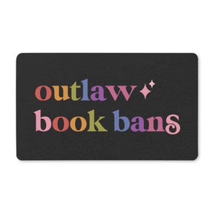May include: A rectangular black doormat featuring the text "outlaw book bans" in a multi-colored serif font. The letters are colored in shades of pink, yellow, blue, purple, and orange. A small four-pointed star icon is positioned to the right.