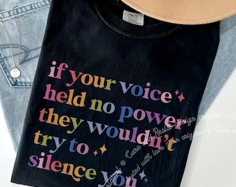 Może przedstawiać: Czarny t-shirt z napisem "if your voice held no power they wouldn't try to silence you" w tęczowych kolorach. Koszulka jest połączona z niebieskimi dżinsami i beżowym kapeluszem.