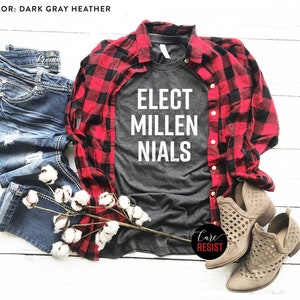 May include: A dark gray heather t-shirt with the text "ELECT MILLENNIALS" in white. The shirt is styled with a red and black plaid shirt, blue jeans, and brown cut-out ankle boots.