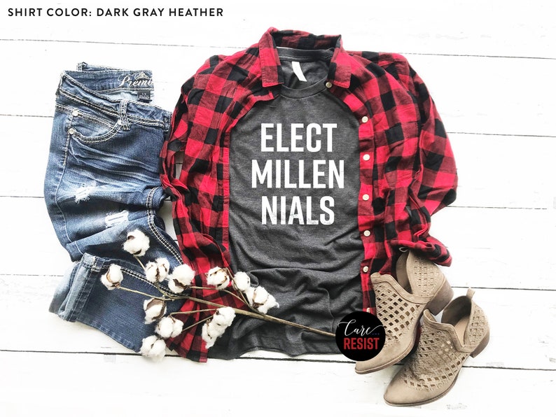May include: A dark gray heather t-shirt with the text "ELECT MILLENNIALS" in white. The shirt is styled with a red and black plaid shirt, blue jeans, and brown cut-out ankle boots.
