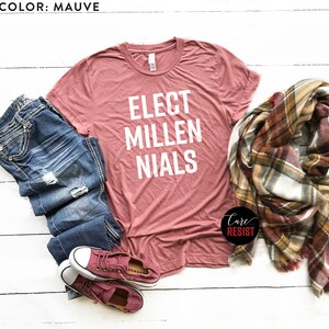 May include: Mauve t-shirt with the text "Elect Millennials" in white lettering. The shirt is laid out on a white wooden surface with a pair of blue jeans, a plaid scarf, and a pair of pink sneakers.