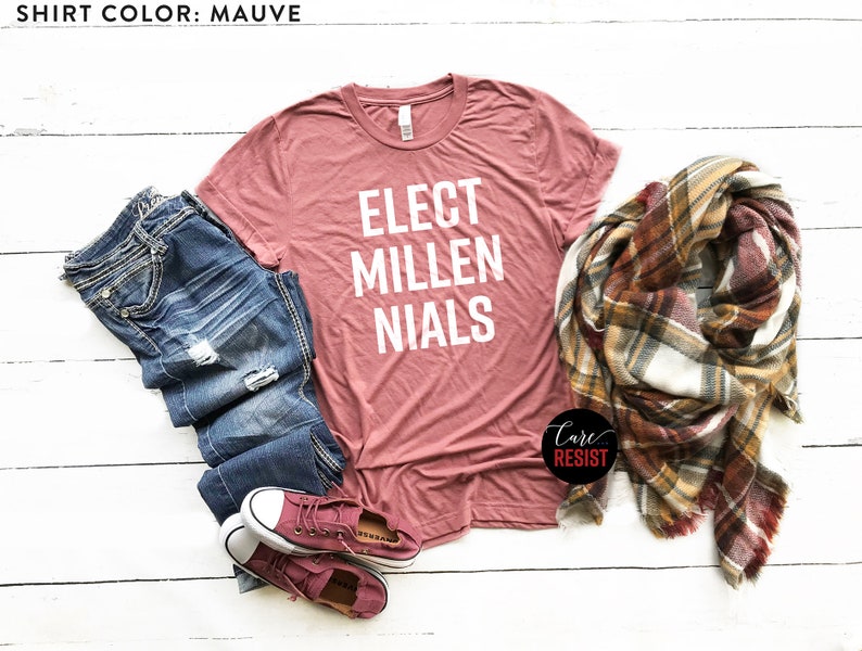 May include: Mauve t-shirt with the text "Elect Millennials" in white lettering. The shirt is laid out on a white wooden surface with a pair of blue jeans, a plaid scarf, and a pair of pink sneakers.
