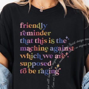 Op de afbeelding: Zwart T-shirt met een regenboogkleurige afbeelding met de tekst "friendly reminder that this is the machine against which we are supposed to be raging".