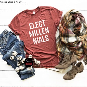 May include: A rust-colored t-shirt with the text "ELECT MILLENIALS" in white lettering. The shirt is laid out on a white wooden surface with a pair of blue jeans, a plaid scarf, and brown ankle boots.