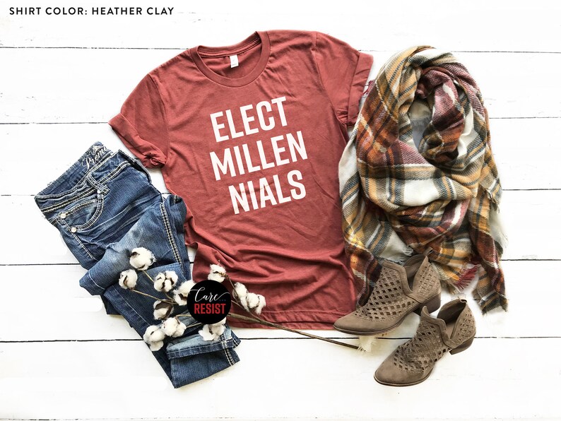 May include: A rust-colored t-shirt with the text "ELECT MILLENIALS" in white lettering. The shirt is laid out on a white wooden surface with a pair of blue jeans, a plaid scarf, and brown ankle boots.